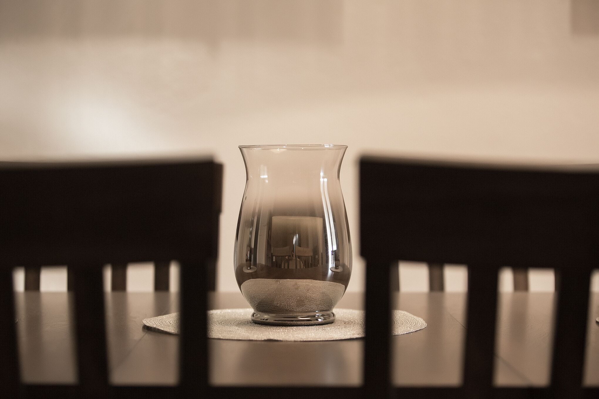 Eine transparente Glasvase auf einem Tisch mit Untersetzer aus gewebtem Material, umgeben von dunklen Holzsesseln.