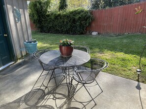 Eine Terrasse mit einem runden Metalltisch und Stühlen, einem Blumentopf und einem blauen Mülleimer.