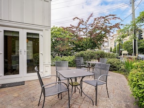 Une terrasse avec des meubles d’extérieur, un bâtiment blanc doté de portes vitrées et un arbre aux feuilles rouges.