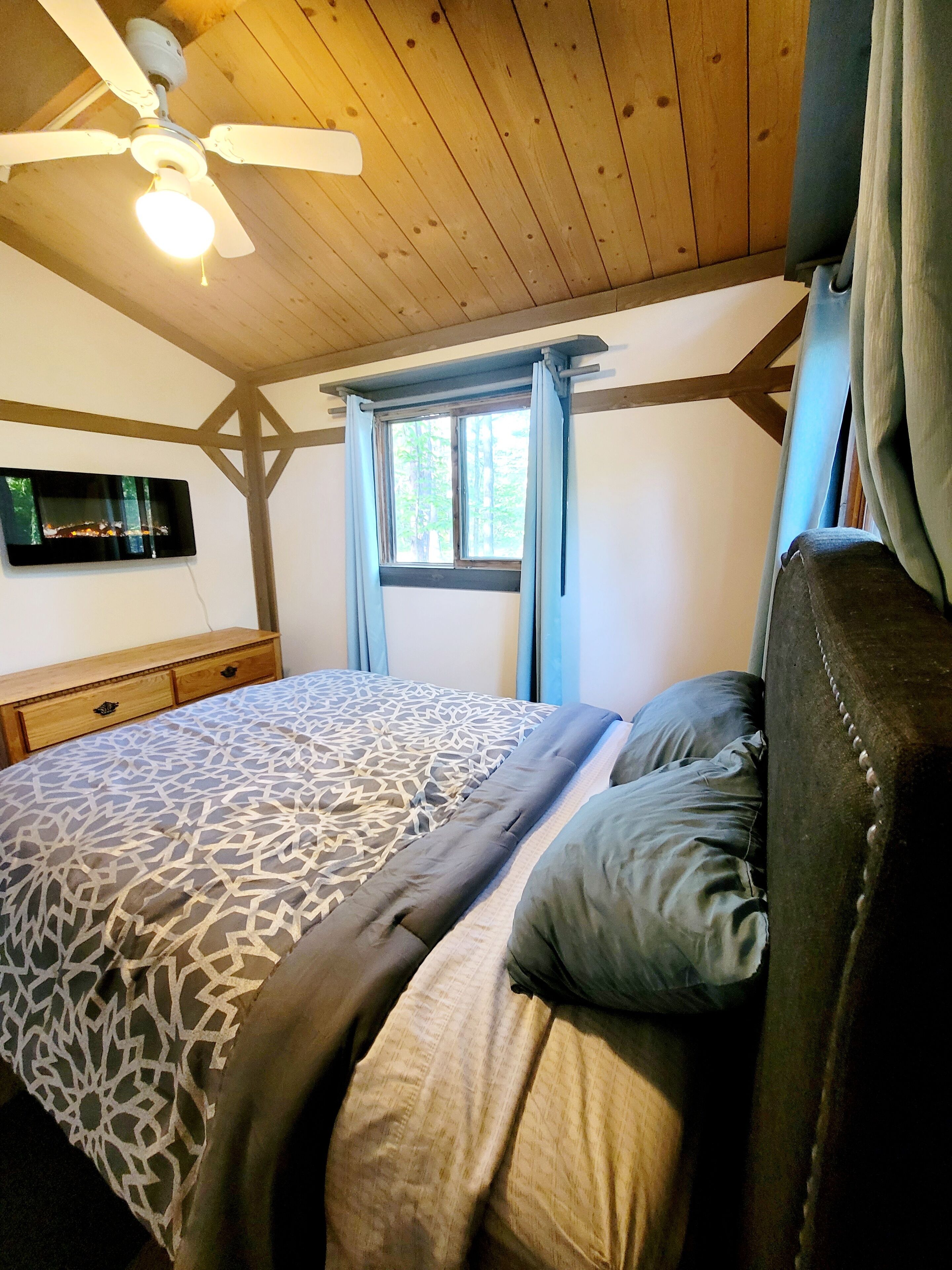 Une chambre à coucher avec un lit, un ventilateur de plafond, un téléviseur fixé au mur et une fenêtre avec des rideaux.