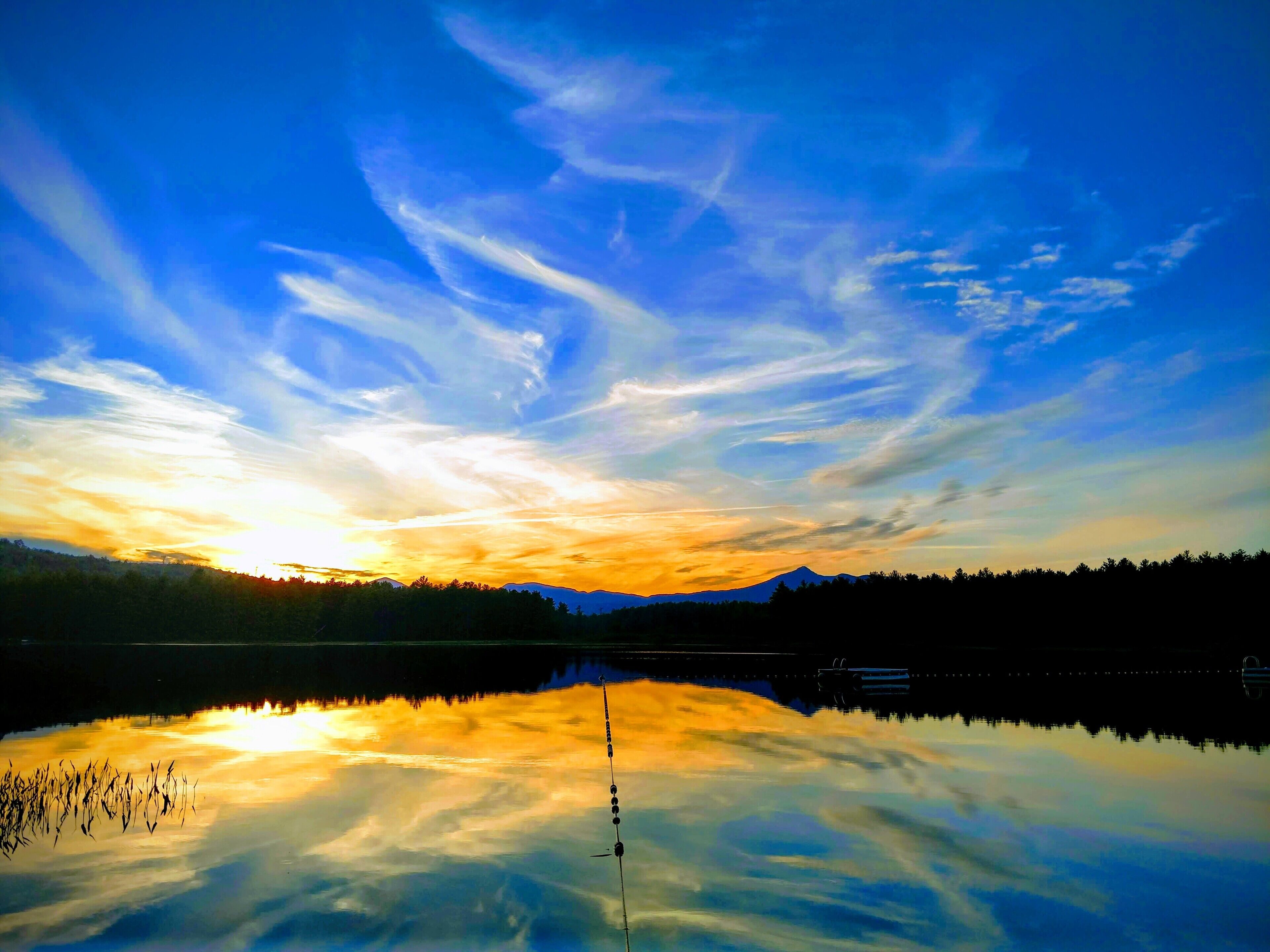 Un lac paisible reflétant le ciel au coucher du soleil et les nuages, entouré d’une zone boisée et dominé par des montagnes lointaines.
