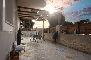 An outdoor patio with stone flooring, a wooden table and chairs, potted plants, and a wooden fence. - Liz Apartment  by CorfuEscapes (Corfu)
