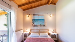 A bedroom with a wooden ceiling, a bed with a pink quilt, bedside lamps, a nightstand, a window with a view of the sea, and a painting above the bed. -  Villa Pine Forest  (Corfu)