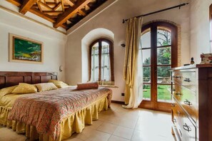 A bedroom with a large bed, a wooden headboard, a painting on the wall, a window with curtains, and a view of the outdoors. - Casa Conti di Sotto (Gavorrano)