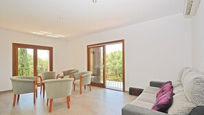 Ein geräumiges Wohnzimmer mit einem grauen Sofa, zwei Sesseln und einem Couchtisch. Große Fenster bieten Blick auf viel Grün draußen.