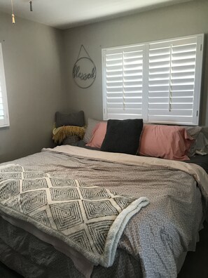 A bedroom with a bed, a window with blinds, a wall decoration, and a hanging light fixture.