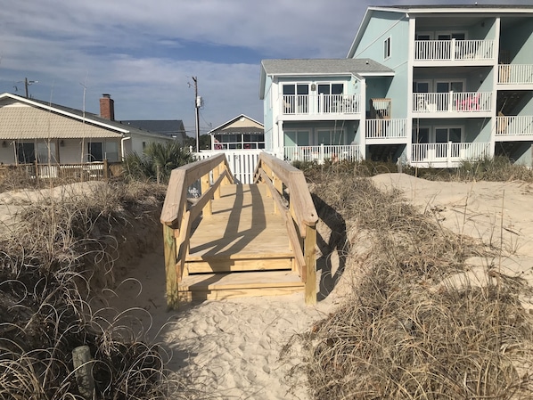Un muelle de madera que conduce a una propiedad frente a la playa, con un edificio de dos pisos y balcones.