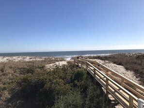 Un malecón de madera que conduce a una playa con dunas y vegetación escasa.