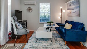 Una sala de estar moderna con un sofá azul, una mesa de centro blanca, un sillón gris y un ventanal con persianas.