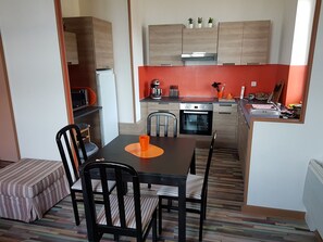 Une cuisine compacte dotée d’appareils électroménagers modernes, un coin repas avec une table et des chaises, et un petit espace détente avec un banc.