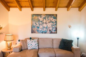 A living room with a beige sofa, a round side table with a lamp, and a painting of a tree on the wall.