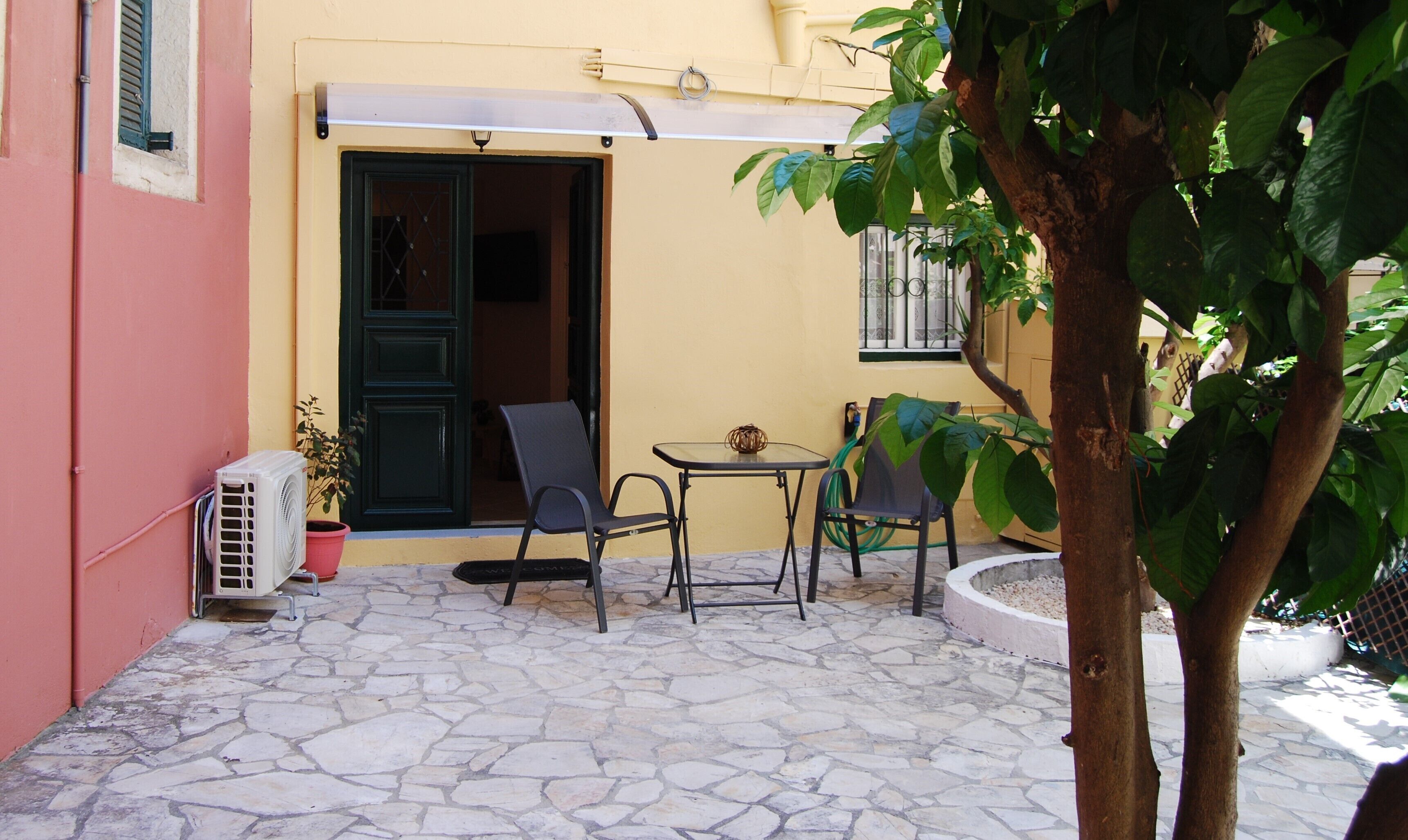 Un pequeño patio al aire libre con suelo de piedra, dos sillas, una mesa, una planta en maceta y la entrada de un edificio con una puerta y una ventana.