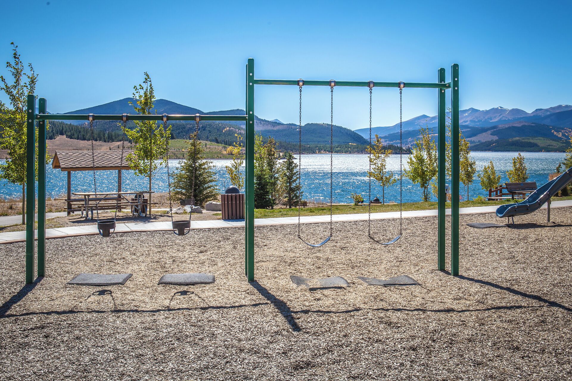 A playground with swings and a slide, picnic tables, and a bench near a lake with mountains in the background.