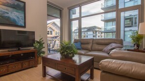 A living room with a television, a wooden coffee table, a sofa, and large windows offering a view of neighboring houses. - Duet Delight-Beautiful Double Ensuite Condo in James Bay, Wonderful Neighbourhood by Victoria Prime! (Victoria)