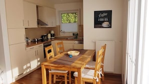 Une cuisine de taille réduite avec une table et des chaises en bois, une décoration murale sur le thème du café et une fenêtre laissant entrer la lumière naturelle.
