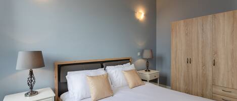A neatly made bed with white and beige pillows, a wooden headboard, and a nightstand with a lamp. There is a wardrobe with two doors and a small drawer.