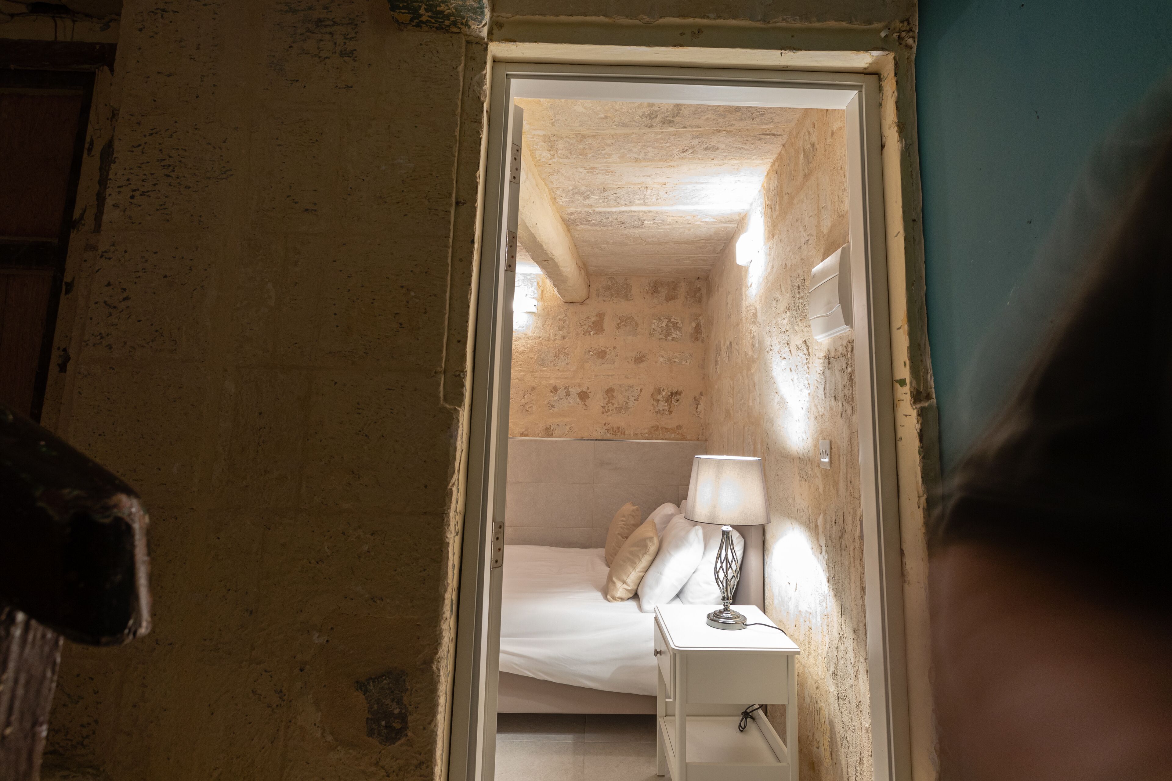 A narrow hallway leading to a small, dimly lit room with a bed, a nightstand, and a lamp.