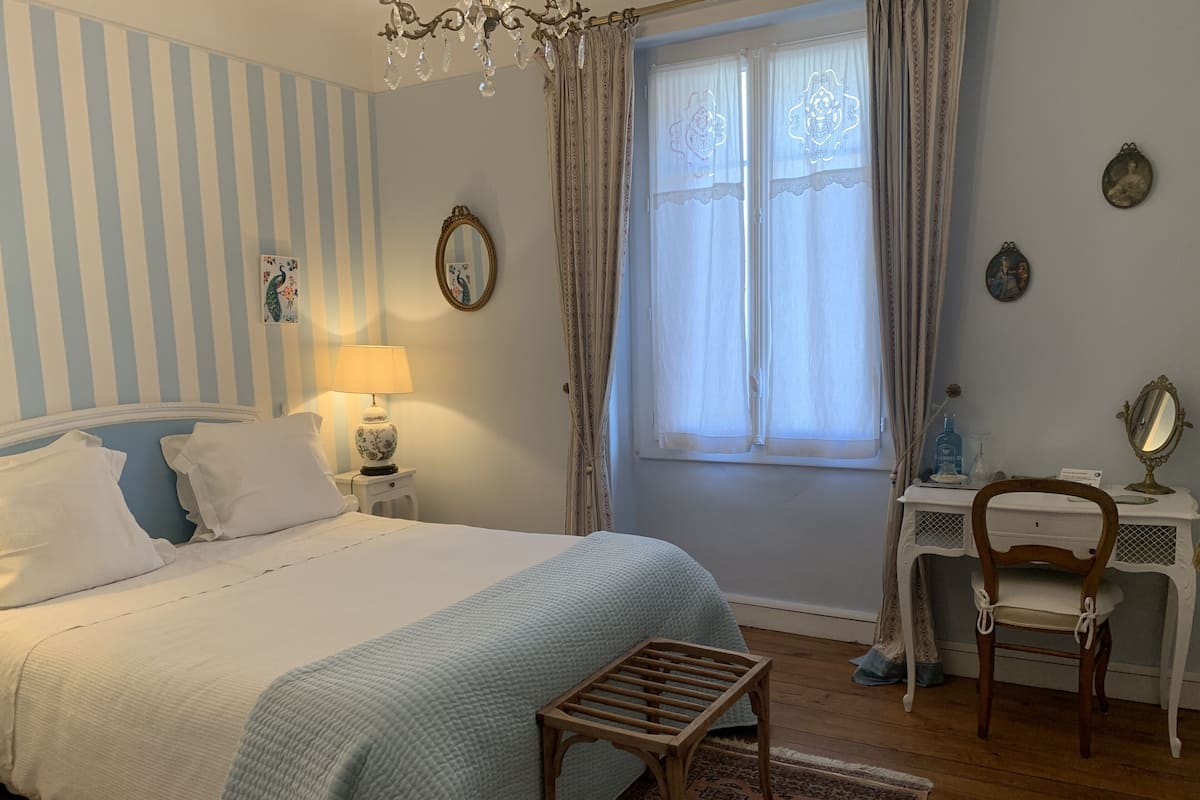 A bedroom with a bed, a nightstand with a lamp, a dresser with a mirror, a chair, and a window with curtains.