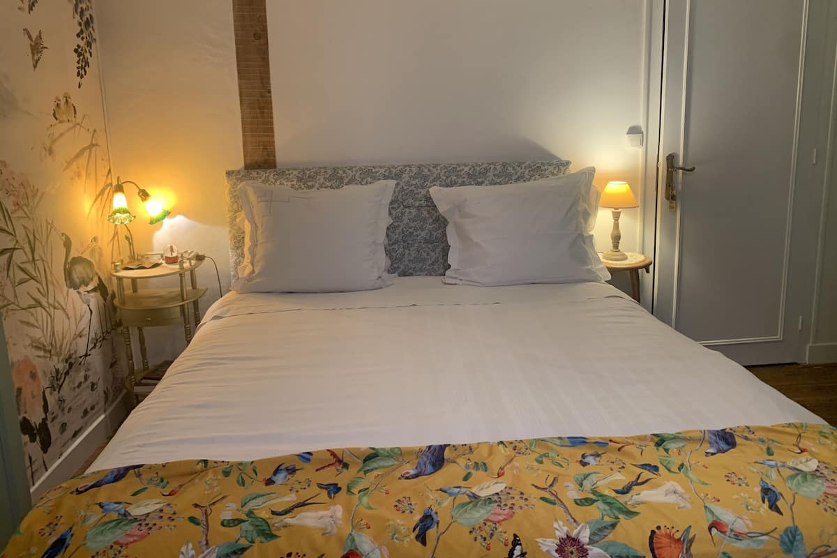 A neatly made bed with white linens, a floral-patterned bedspread, and two white pillows. There is a nightstand with a lamp and a small table beside the bed. The room has a wooden headboard and a door to the right.