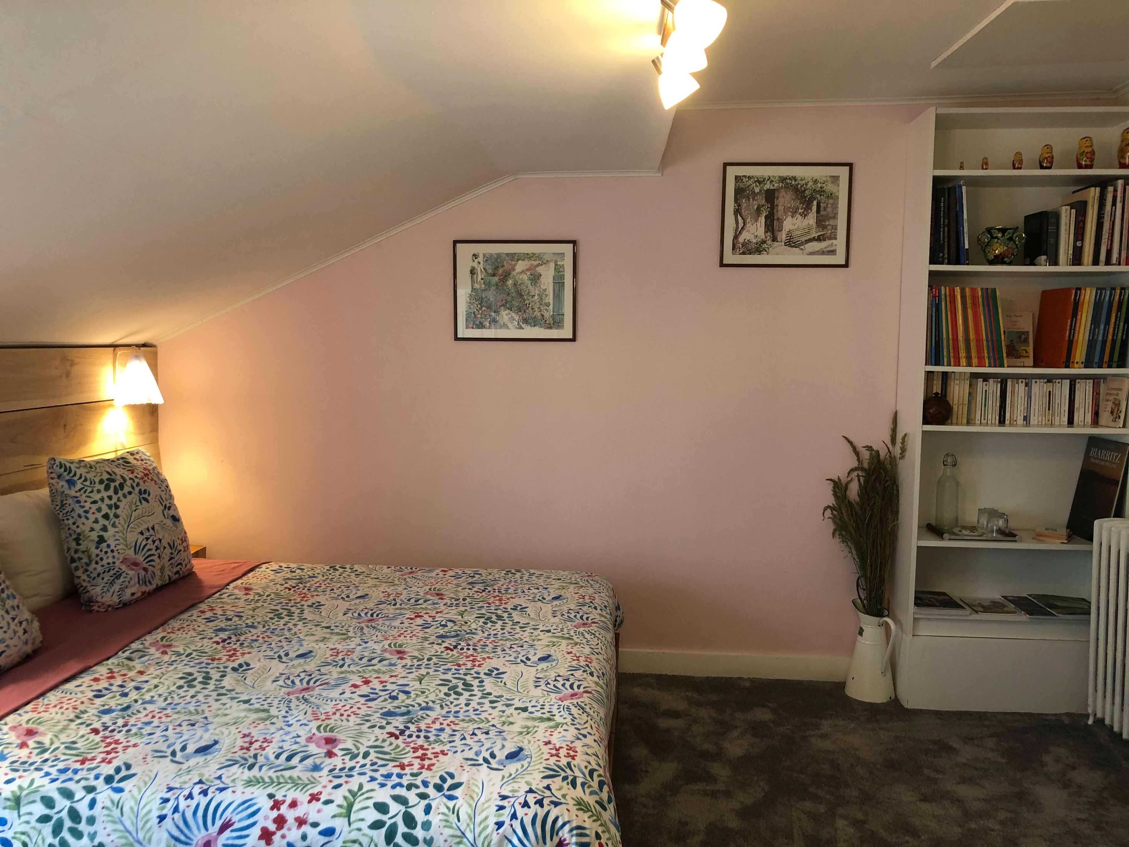A bedroom with a floral bedspread, a bookshelf with books, and two framed pictures on the wall.