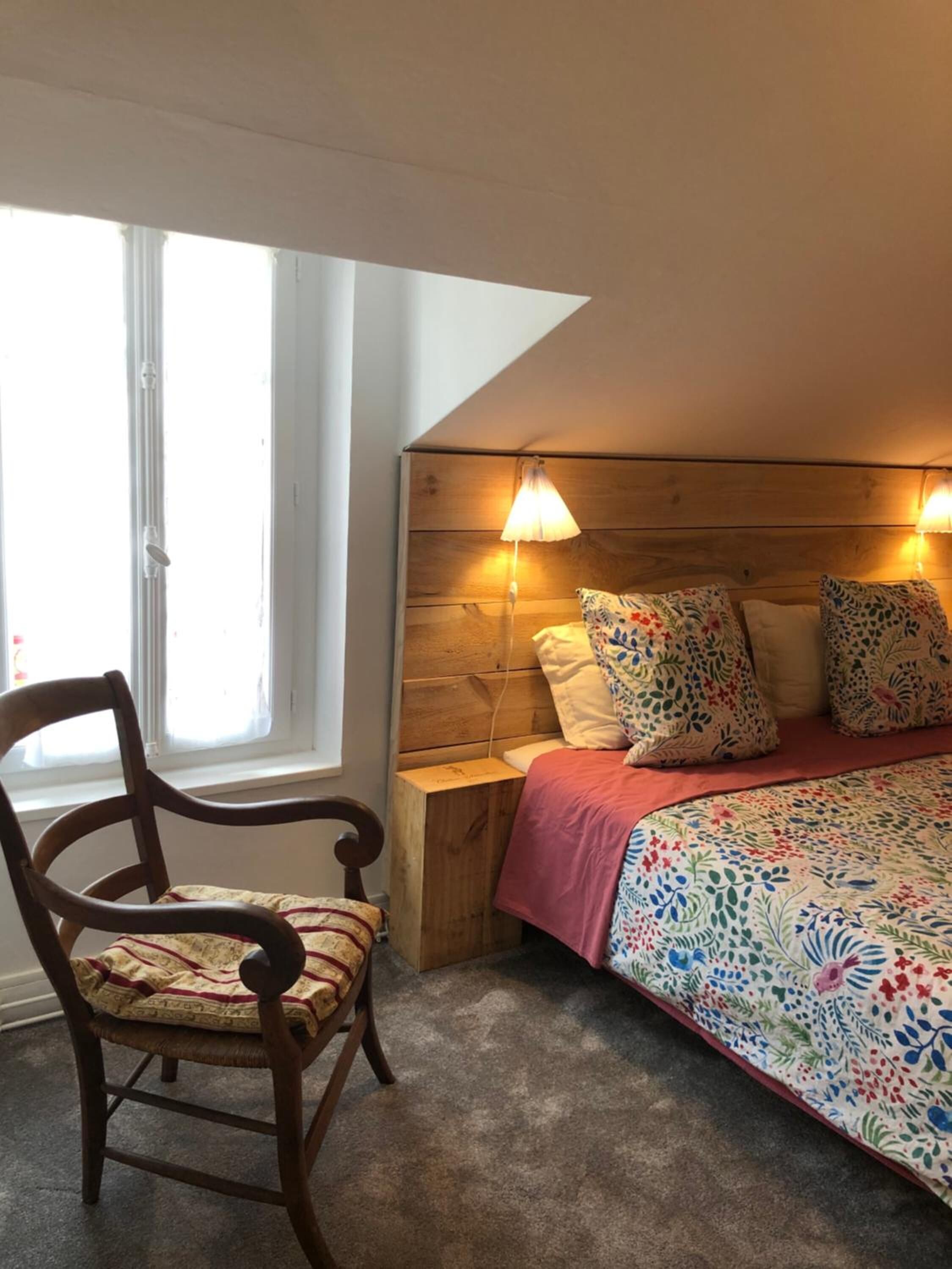 Une chambre avec un plafond en pente, une tête de lit en bois, un lit avec une literie à motifs floraux, une table de chevet, une chaise et une fenêtre avec des rideaux.