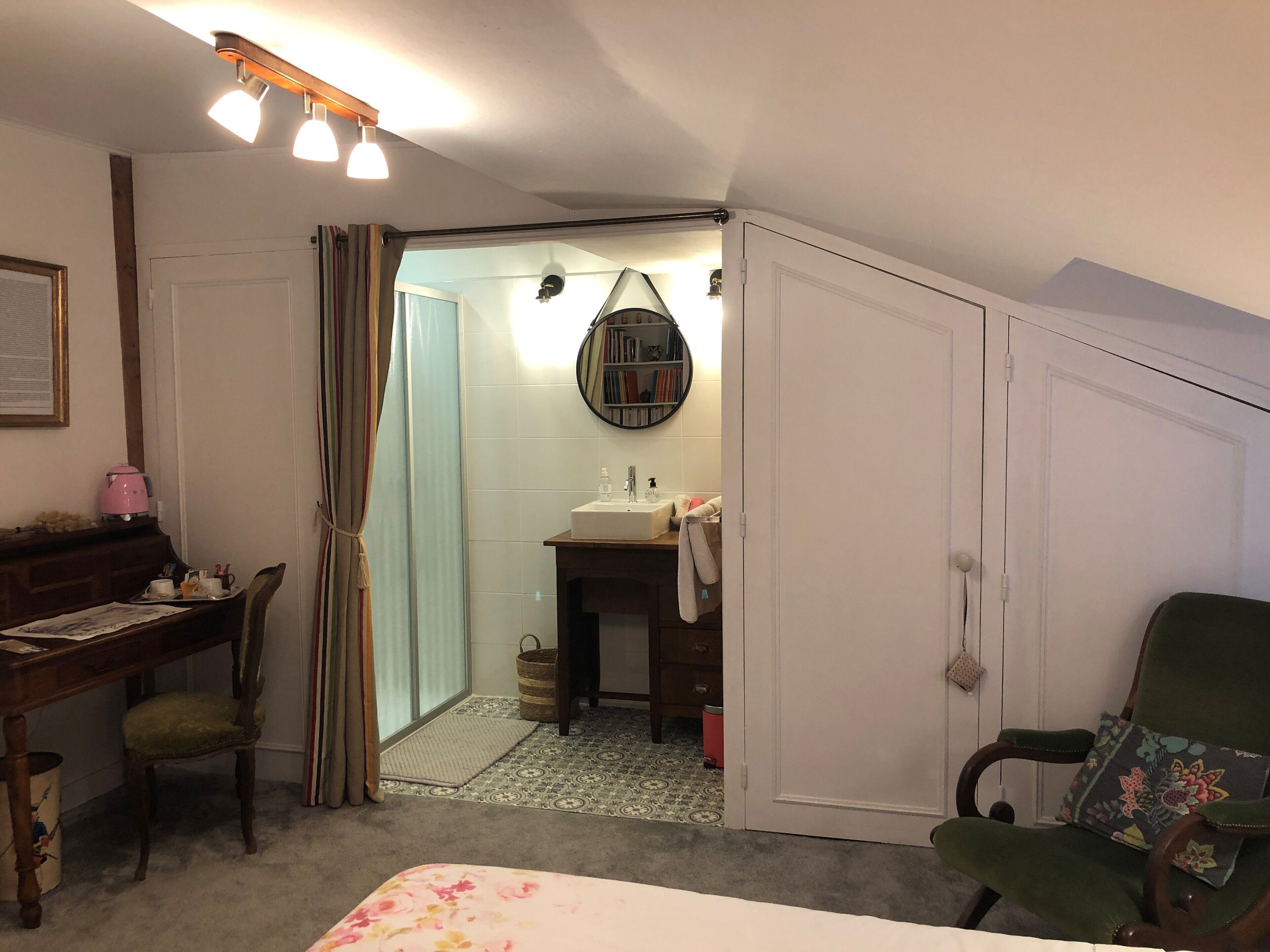 A bedroom with a bed, a desk, a chair, a wardrobe, and a bathroom with a sink and mirror.