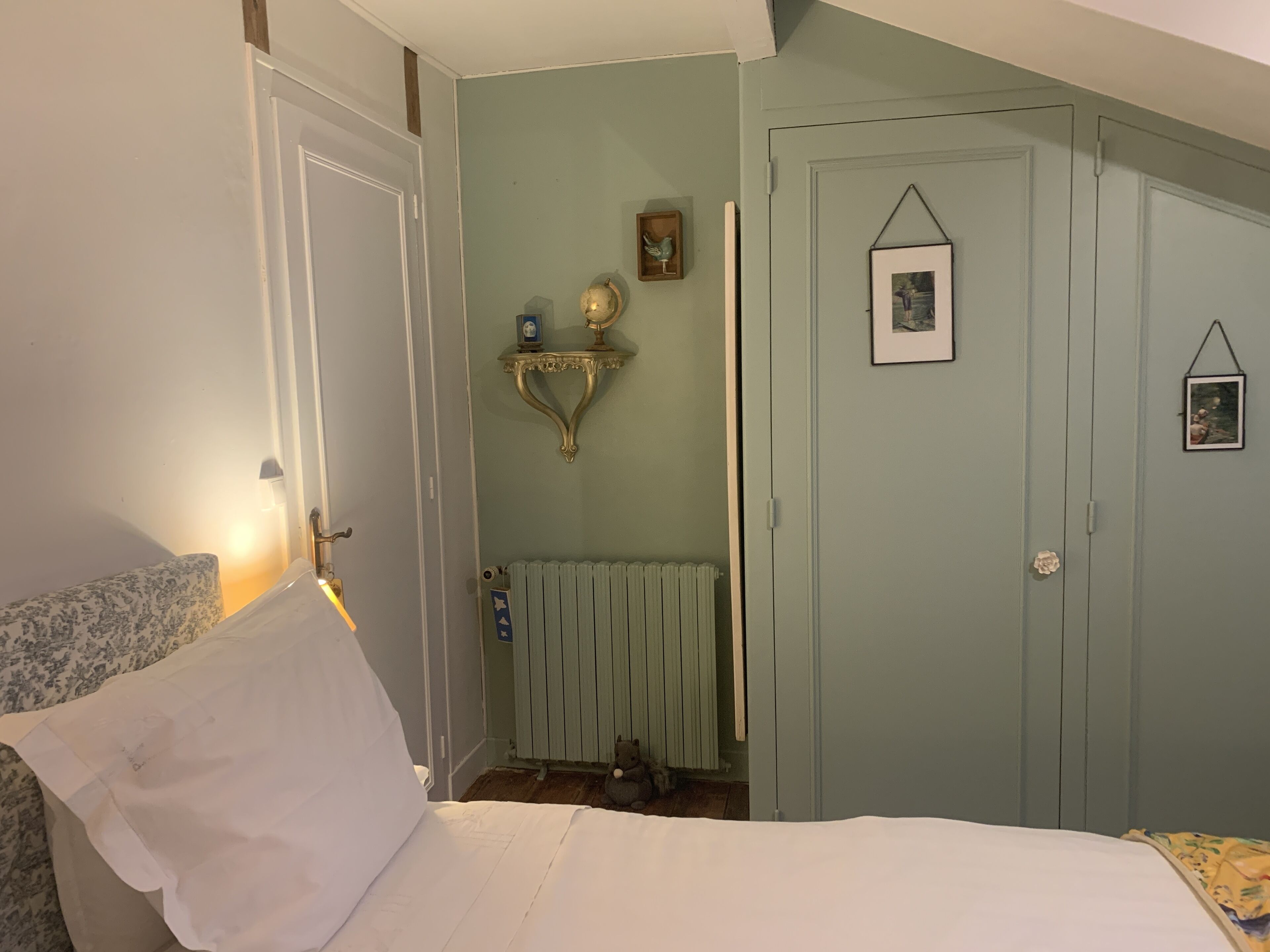A bedroom with a bed, a radiator, a small shelf with decorative items, and a door with a picture frame.