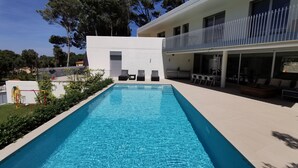 Un espace piscine moderne avec une eau d’un bleu limpide, un bâtiment blanc et un balcon agrémenté de mobilier d’extérieur.