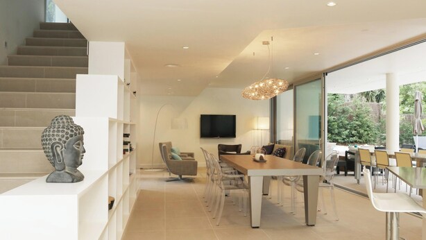 Un intérieur moderne avec un escalier, un coin repas comprenant une longue table et des chaises, et une grande fenêtre donnant sur l’extérieur.