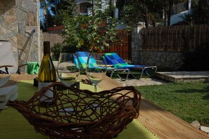 A wooden table with a woven basket, a bottle of wine, and two glasses, set outdoors with a stone wall and a wooden fence in the background.