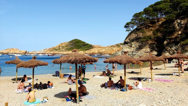 Ein Strand mit Menschen, die sonnenbaden und schwimmen, Reetdachschirmen und Booten im Wasser.