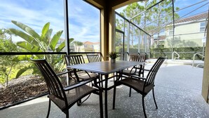 Une terrasse couverte avec une table et des chaises en bois, de grandes fenêtres offrant une vue sur un quartier résidentiel et des arbres.