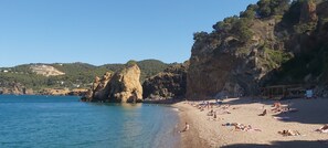 A beach with people sunbathing, a rocky cliff, and a building in the background. - Casa de la Luz luxurious villa by the sea (Begur)