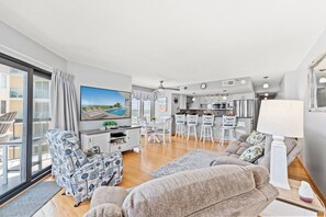 A modern living room with a sofa, a dining area with a table and chairs, a kitchen with a refrigerator and a bar area, and a large window with a view of a pool. - Updated!!! Crescent Towers II 708,  2 BR Ocean Front Luxury Condo with Pool (North Myrtle Beach)