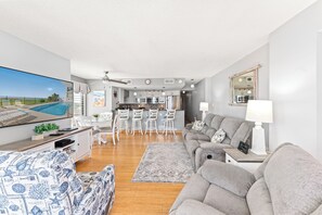 A modern living room with a large flat-screen TV, a grey sofa, a patterned armchair, and a dining area with white bar stools.