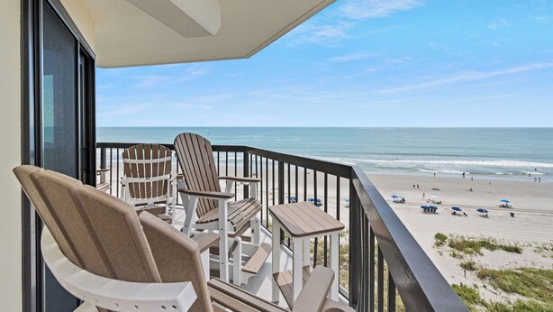 A balcony with Adirondack chairs and a small table, overlooking a beach with people and umbrellas. - Updated!!! Crescent Towers II 708,  2 BR Ocean Front Luxury Condo with Pool (North Myrtle Beach)