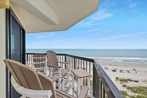 Un balcon avec des fauteuils Adirondack et une petite table, donnant sur une plage où l’on aperçoit des gens et des parasols.