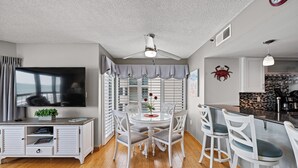 A modern living room with a TV, dining table, and chairs. There is a kitchen area with a bar and a red crab decoration on the wall. - Updated!!! Crescent Towers II 708,  2 BR Ocean Front Luxury Condo with Pool (North Myrtle Beach)