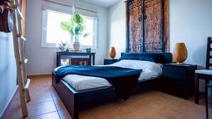 Een slaapkamer met een bed, nachtkastjes, een raam met een plant en een houten deur.