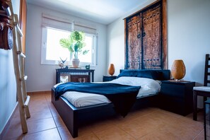 Une chambre à coucher avec un lit, une table de chevet, une lampe, une fenêtre avec des stores et une plante en pot.