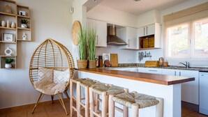 Een keuken met een houten eiland, barkrukken, een rieten stoel en een plank met decoratieve objecten.