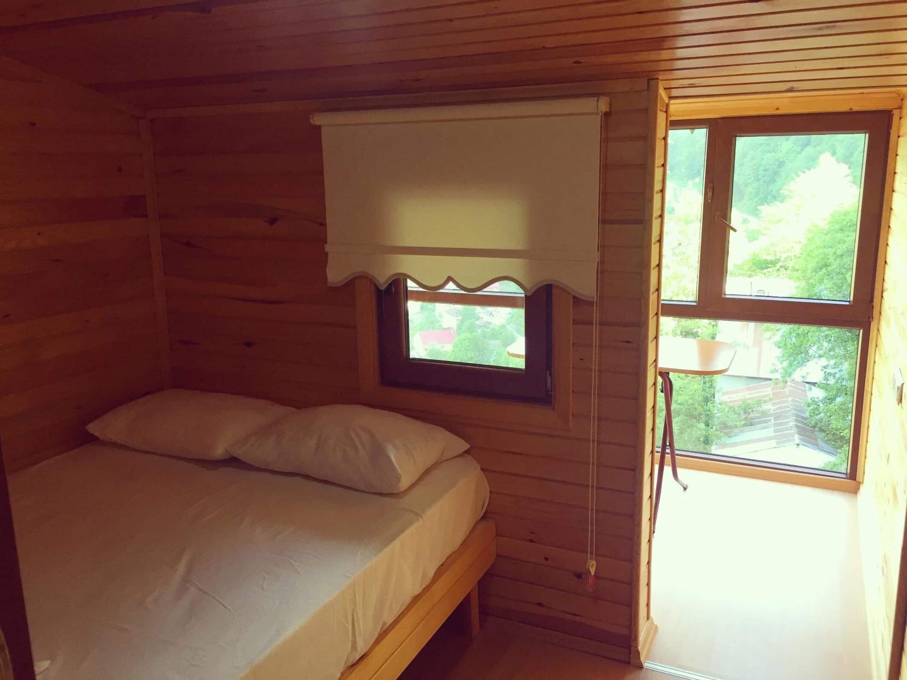Habitación de cabaña de madera con una cama individual, una ventana con cortinilla blanca y vistas a la vegetación.
