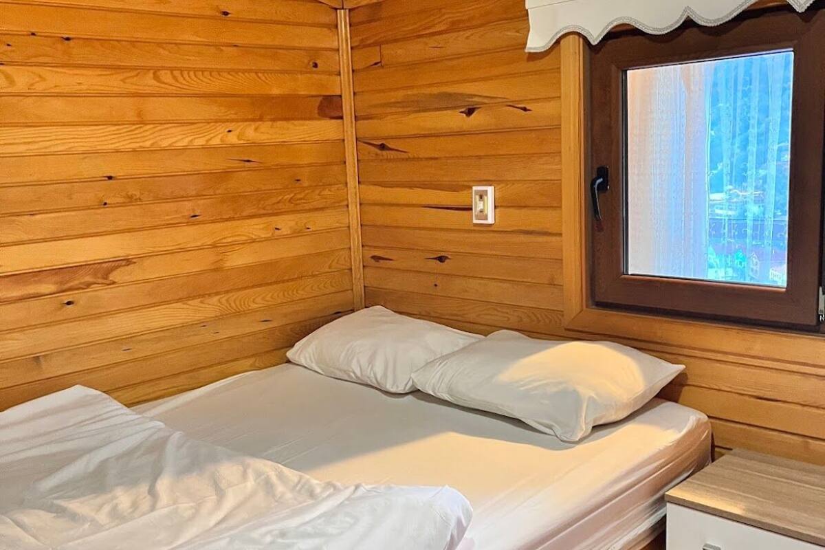 A small cabin with two single beds, a wooden ceiling, a window with a view of trees, and a small white desk.