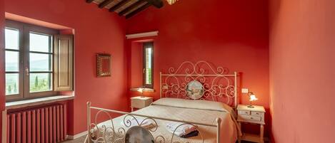 A bedroom with a red wall, a wooden ceiling, a metal bed frame, a bedside table with a lamp, and a window with a view.