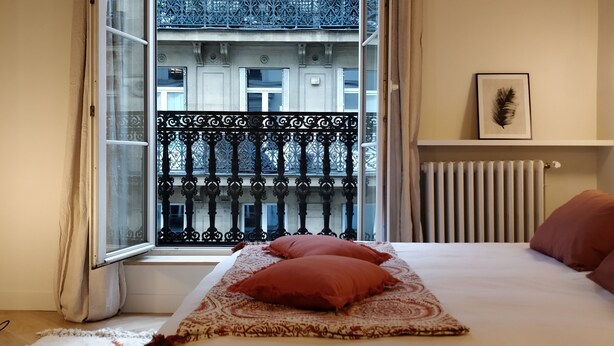 Un dormitorio con una cama grande, una ventana con vistas a un edificio, un radiador y un cuadro en la pared.