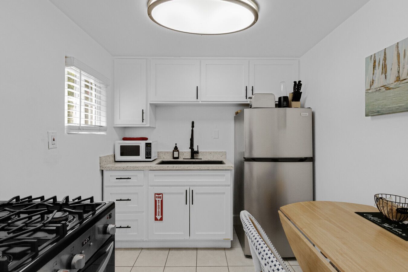 A modern kitchen with white cabinets, a stainless steel refrigerator, a microwave, a gas stove, and a wooden dining table.