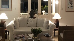 A living room with a white sofa, a coffee table, and two lamps. There are framed pictures on the walls and a window with a view of the outside. - Captivating 3-bed Villa in Montego Bay (Montego Bay)