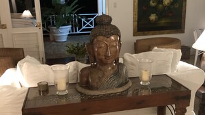A room with a Buddha statue on a glass table, white cushions, a painting, and a view of the outside. - Captivating 3-bed Villa in Montego Bay (Montego Bay)