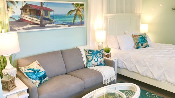 Une pièce comprenant un lit, un canapé, une table basse avec un plateau en verre, un tableau représentant une scène de plage et une décoration murale en forme d’étoile de mer.