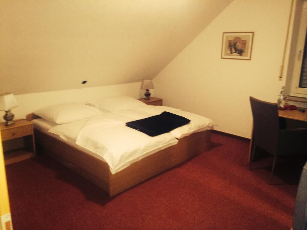 Una cama individual con ropa de cama blanca, una mesita de noche pequeña con una lámpara, una silla e imagen enmarcada en la pared.
